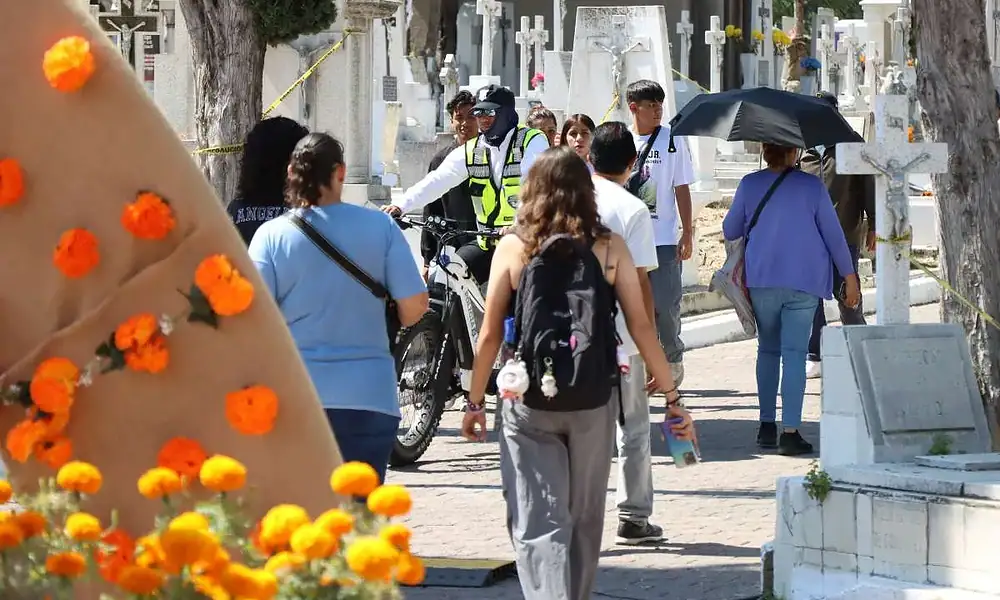 Personas caminando en cementerio durante el día, con decoración de flores naranjas y cruces blancas.